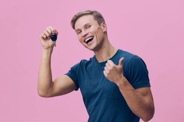 Kısa kahverengi saçlı, günlük mavi tişörtlü mutlu bir adam, araba anahtarı tutarken geniş bir gülümseme, baş parmak hareketi, pembe arka planda izole edilmiş, başarı, heyecan ve