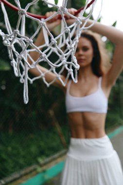 Dinamik bir sahnede beyaz etekli, atletik bir kadın basketbol potasına karşı poz veriyor, yemyeşil bir arka planda güç ve enerji yayıyor.