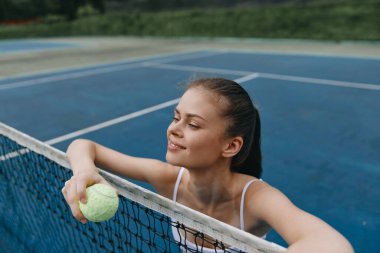 Genç bir bayan sporcu, elinde parlak sarı bir tenis topuyla tenis ağına yaslanıyor. Yüzündeki ifade mutluluk ve kararlılık yayıyor. Canlı bir mavi tenis kortunun karşısında duruyor.