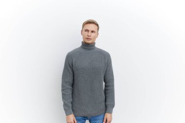 Sarı saçlı, rahat gri bir balıkçı yaka kazak giyen, kameraya özgüvenli bir şekilde bakan, beyaz arka plan, günlük tarz, moda portresi, erkek yaşam tarzı