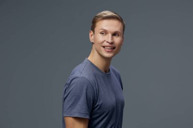 Gri tişörtlü, dizüstü bilgisayarla çalışan gülümseyen bir adam düşünceli, genç, beyaz, dijital göçebe uzaktan kumanda konseptini bir yana bırakıp karanlık stüdyo arka planına bakıyor.