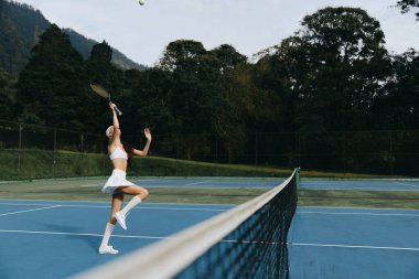 Beyaz giyinmiş neşeli bir bayan tenisçi, sahada dinamik bir poz veriyor, yemyeşil ve mavi gökyüzü ile çevrili yaz sporları temaları için mükemmel.