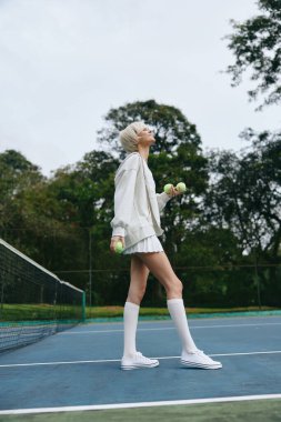 Enerjik genç bayan sporcu şık spor kıyafetleriyle bir tenis kortunun üzerinde ağırlık tutuyor, yeşillik ile çevrili, sağlık, zindelik ve kararlılık temaları iletiyor.