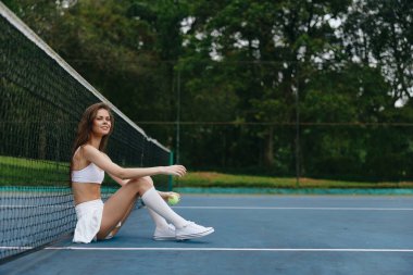 Spor kıyafetleri giymiş genç bir kadın tenis kortunda oturuyor, neşeyle gülümsüyor ve elinde bir tenis topu tutuyor yemyeşil arka plan canlı açık hava sahnesine canlılık katıyor.