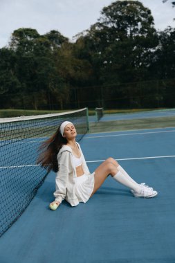 Beyaz spor kıyafetleri giymiş genç bir kadın tenis kortunda oturuyor, doğal ortamda mutluluk ve sükunet yayıyor, sportif bir yaşam tarzını somutlaştırıyor.