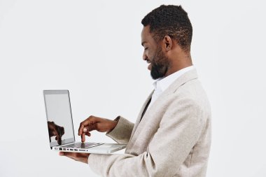 Koyu tenli, açık bej takım elbiseli, dizüstü bilgisayarla uğraşan, beyaz arka planda duran kendinden emin bir adam. Teknoloji, uzaktan çalışma, profesyonellik, modern
