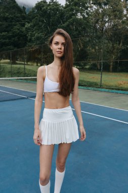 Beyaz etek ve spor sutyen giyen kendine güvenen genç bayan tenisçi, yemyeşil ile çevrili bir kortta atletizm sergiliyor.