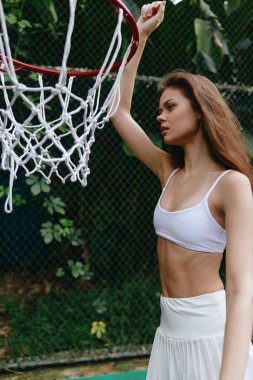 Beyaz spor kıyafetleri içinde kendine güvenen, açık havada basketbol oynayan sporcu, atletizm ve canlı yeşil bir geçmişi olan, aktif bir yaşam tarzını teşvik eden genç bir kadın.