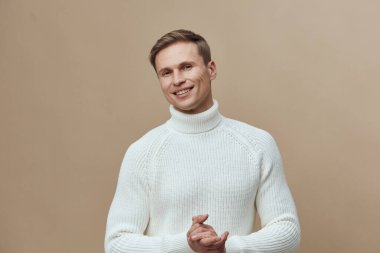 Kahverengi arka planda izole edilmiş beyaz örülü balıkçı yaka kazak giyen gülümseyen genç beyaz adam kameraya sakin ve rahat yüz ifadesine zihinsel sağlık ve aletler kavramına bakıyordu.