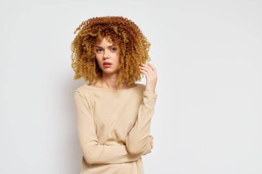 Güzel kıvırcık saçlı bir kadın bej bir bluz giyen düz beyaz bir arkaplanda poz verir. Kendine güveni ve yumuşak ifadesi doğal buklelerin güzelliğini ön plana çıkarır.