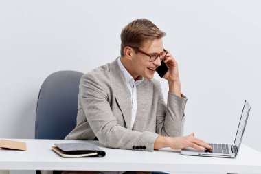 Sırıtkan, beyaz gözlüklü, rahat ceketli, akıllı telefondan konuşan, dizüstü bilgisayarda beyaz arka planda yazan genç beyaz bir adam. Dijital aygıtlar kullanarak birden fazla iş yapan iş adamı