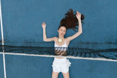 Modayı takip eden beyaz kıyafetli enerjik genç bir kadın, bir tenis topuyla neşe içinde etkileşim içinde, yaz titreşimleri yayıyor ve çarpıcı bir mavi zemine karşı coşkulu.