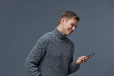 Sade gri örgü süveterli, kısa saçlı mutlu bir adam gri arka planda gülümseyen akıllı telefona bakıyor. Dijital iletişimin keyfi, rahat yaşam tarzı, modern teknoloji.