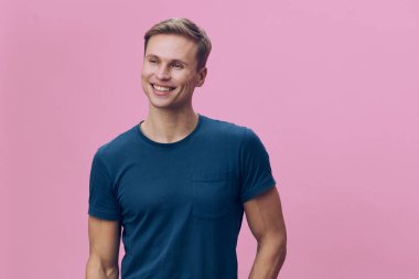 Kısa kahverengi saçlı, günlük mavi tişörtlü neşeli genç adam, içten gülümseyen ve kameraya bakan, pastel pembe arka planda duran, mutluluğu vurgulayan bir portre, özgüven