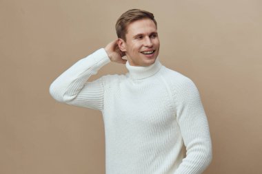 Beyaz örgülü balıkçı yaka kazak giyen gülümseyen genç beyaz bir adam boynunun arkasına rahatça dokunuyor ve kapalı arka planda izole edilmiş mutlu bir ifadeyle yüzünü çeviriyor.