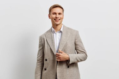 Neşeli gülümseyen, zarif açık gri takım elbiseli, beyaz gömlekli, göğsünde elle poz veren, sade beyaz stüdyo geçmişi olan, profesyonel iş kıyafeti, pozitif ruh hali olan genç bir adam.