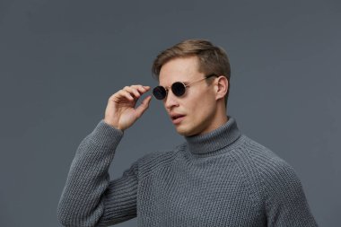 Kısa kahverengi saçlı, açık tenli, gri balıkçı yaka kazak ve güneş gözlüğü takan, tarafsız bir arkaplan önünde rahatça poz veren şık bir adam. Moda, zarafet, güven konsepti