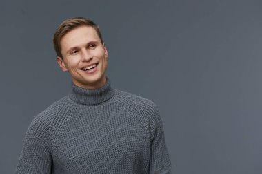 Gri balıkçı yaka kazak giyen mutlu genç bir adam stüdyo ortamında nötr gri bir arkaplanda dikilirken ışıl ışıl gülümsüyor. Görüntü neşeli, rahat bir ruh hali yakalıyor.