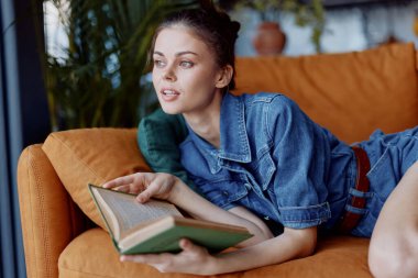 Evde oturup kitap okuyan bir kadın rahat oturma odasında kot gömleğiyle kitap okuyor.