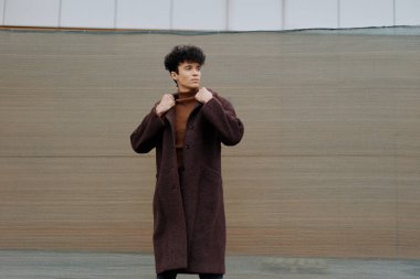 Kahverengi ceketli moda adamı beton zeminde elleri kalçasında ve ceplerinde duruyor..