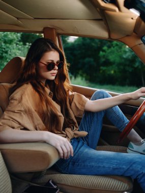 Modaya uygun genç bir kadın klasik bir arabada oturuyor, tarzını güneş gözlüğü ve rahat, rahat kıyafetlerle sergiliyor, canlı yeşilliklerle çevrili, yaz temaları için mükemmel..