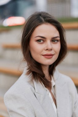 Kendine güvenen, bej ceketli, hoş gülüşlü, şehir arka planı bulanık, doğal ışık, tarafsız tonlar, moda konsepti olan, açık havada oturan bir kadın.