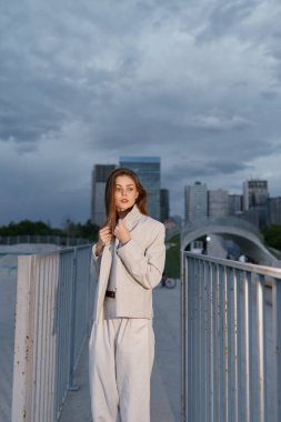 Açık bej takım elbiseli kendine güvenen bir kadın gökdelenleri ve bulutlu gökyüzü olan metal bir şehir köprüsünde duruyor. Modern iş modasını ve sonbahar havasını simgeliyor..