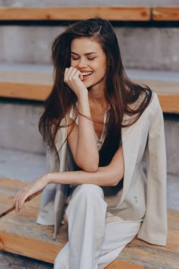 Bej renkli takım elbiseli genç bir kadın, tahta merdivenlerde otururken kendine güvenerek gülümsüyor. Parlak, doğal bir ışıkla. İlkbaharda gündelik moda ve pozitif duygular sergiliyor..