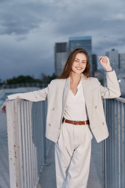 Açık bej takım elbise ve beyaz bluz giyen kendine güvenen bir kadın bulutlu gökyüzünün altında şehir köprüsünde rahatça duruyor ve başarısını ve modern iş tarzı tarzını ifade ediyor.