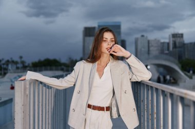 Bej renkli iş elbisesi giymiş şık bir kadın, şehir siluetine karşı poz veriyor, sakin, düşünceli bir ifadeyle özgüveni yansıtıyor, kurumsal temalar için ideal..