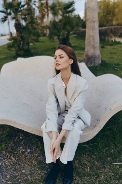 Açık gri takım elbiseli şık bir kadın baharda yemyeşil ağaçlı şık bir açık hava bankında oturarak modern iş tarzını ve sakin havasını sergiliyor.