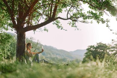 Genç bir kadın zarif bir şekilde bir ağacın altında oturmuş yemyeşil bir içeceği yudumlarken manzara dinginliği ve sağlıklı bir yaşam tarzını yansıtıyor.