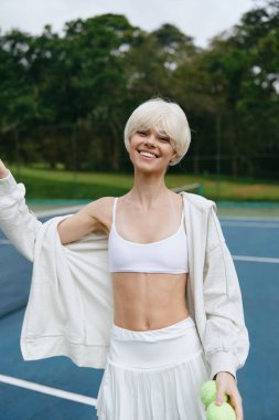 Kısa sarı saçlı enerjik genç bir kadın beyaz spor bluz ve etek giyiyor, tenis kortunda neşe içinde poz veriyor, fitness ve sağlıklı bir yaşam tarzını temsil ediyor canlı yeşil bir arkaplanın ortasında