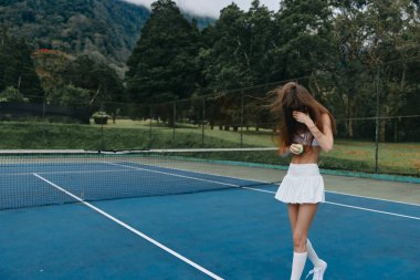 Genç bir tenisçi, mavi bir kortta beyaz bir elbiseyle, yeşil bir manzara ve dağların arasında, neşeyle bir top tutuyor, oyunun ve yazın ruhunu yakalıyor.