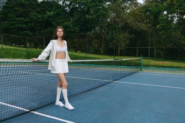 Beyaz spor elbiseli, kendine güvenen genç bir kadın tenis ağının yanında poz veriyor, sağlıklı bir yaşam tarzı ve spor hevesi sergiliyor, fitness ve spor temaları için ideal.