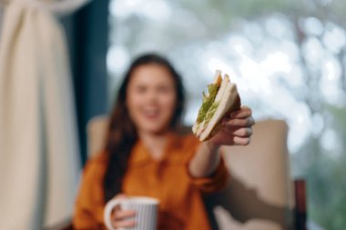 Uzun saçlı genç bir kadın, elinde lezzetli bir sandviç ve kahve fincanı tutarken gülümsüyor. Sıcak tonlarla sıcak bir yerde kurulmuş.