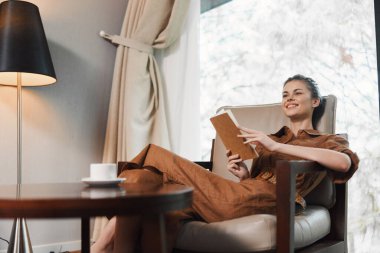 Rahat genç bir kadın, şık kahverengi bir tulum giymiş, modern bir oturma odasında sıcak renk paleti olan bir kitap okurken gülümsüyor.