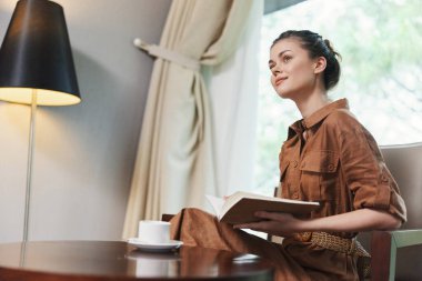 Kahverengi elbiseli genç bir kadın elinde kitapla sessizce oturur, pencerenin kenarında yumuşak, doğal bir ışıkla okumaya dalmış, rahat ve şık bir atmosfer yaratır.