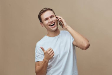 Neşeli, gülümseyen bir adam, cep telefonuyla bir telefon görüşmesi yapıyor. Baş parmak hareketleriyle, günlük kıyafetleriyle, açık kahverengi saçlarıyla, tarafsız bir bej arka planda. Modern yaşam tarzı ve iletişim konsepti