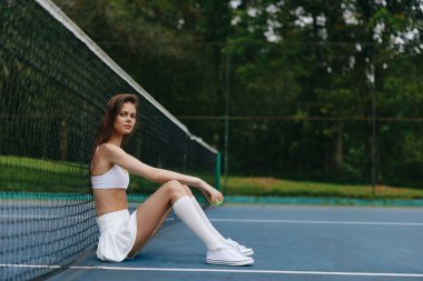 Şık spor kıyafetleri içinde genç bir kadın tenis ağının yanında oturuyor, kendine güven ve rahatlama yayıyor enerjik yeşil arka plan sportif hissi arttırıyor, forma ve yaşam tarzı için mükemmel.