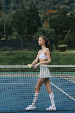 Beyaz spor kıyafetli, kendine güvenen genç bir kadın canlı yeşil bir kortta tenis oynuyor, somut bir atletizm ve neşe, sağlık ve spor kavramları için mükemmel.