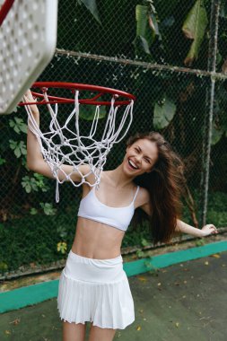 Uzun saçlı, beyaz spor kıyafetleri giyen neşeli genç bayan sporcu canlı yeşil bir ortamda basketbolu seviyor. Spor, spor ve gençlik temaları için mükemmel.
