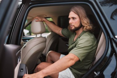 Modern bir arabanın arka koltuğunda oturan bir adam, derin düşünceli bir ifadeyle dışarı bakıyor. İçi açık renkli deri koltuklar içeriyor.