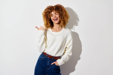 Şehvetli kıvırcık saçlı neşeli bir genç kadın sağını gösterirken geniş bir gülümseme sergiliyor rahat beyaz bir kazak ve yüksek bel kot pantolon giyiyor, el değmemiş beyaz bir arka planda duruyor.