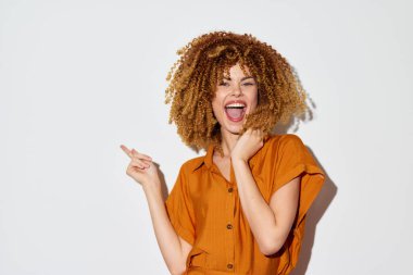 Turuncu elbiseli kıvırcık saçlı heyecanlı genç kadın, gülümsüyor ve temiz beyaz bir arkaplanı işaret ediyor. Bu görüntü mutluluk ve moda ile ilgili kavramlar için ideal neşe ve pozitif titreşimleri yakalıyor.,