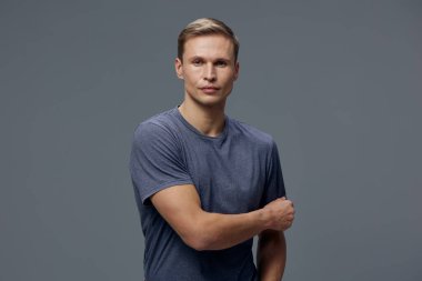 Açık tenli, kısa saçlı, günlük mavi tişörtlü, doğrudan kameraya bakan, dostça bir ifadeyle, düz gri arka planda güvenle poz veren mutlu genç bir adam. Kavram: gündelik biçim