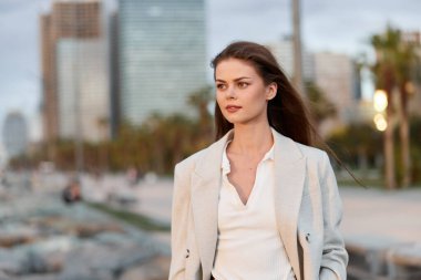 Açık bej ceketli ve beyaz bluzlu kendine güvenen genç kadın şehir silueti ve palmiye ağaçları, doğal makyaj, günlük saç stili, somut modern moda tarzı olan şehir deniz kenarının yanında duruyor.