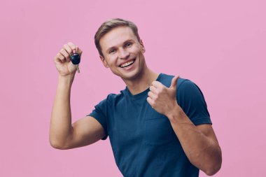 Tüm vücuduyla gülümseyen beyaz bir adam arabanın anahtarlığını tutuyor ve baş parmaklarını kaldırıyor. Günlük mavi tişört giyiyor. İzole edilmiş pembe arka plan stüdyo resmi, dijital göçebe ve serbest çalışma konsepti.