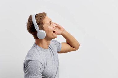 Kısa sarı saçlı neşeli bir kadın, gri tişört giyiyor, büyük kulaklık takıyor, sade beyaz bir arka planda müzik dinlerken mutluluk ve rahatlama yaşıyor.