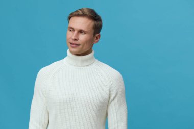 Beyaz balıkçı yaka kazaklı neşeli bir genç adam portresi gülümseyen ve yana bakan, canlı mavi bir arka planda izole. Sıradan moda ve yaşam tarzı fotoğrafı mutluluğu yakalıyor ve...